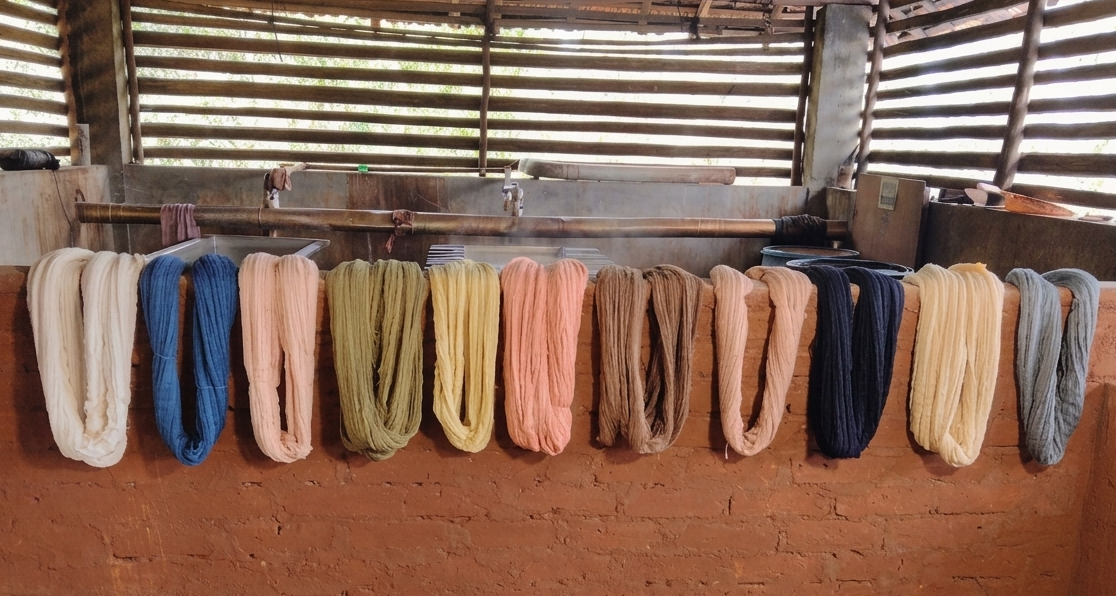 A series of colorful yarn or fabric bales hanging side-by-side on a rustic brick wall inside a workshop with wooden slat windows.